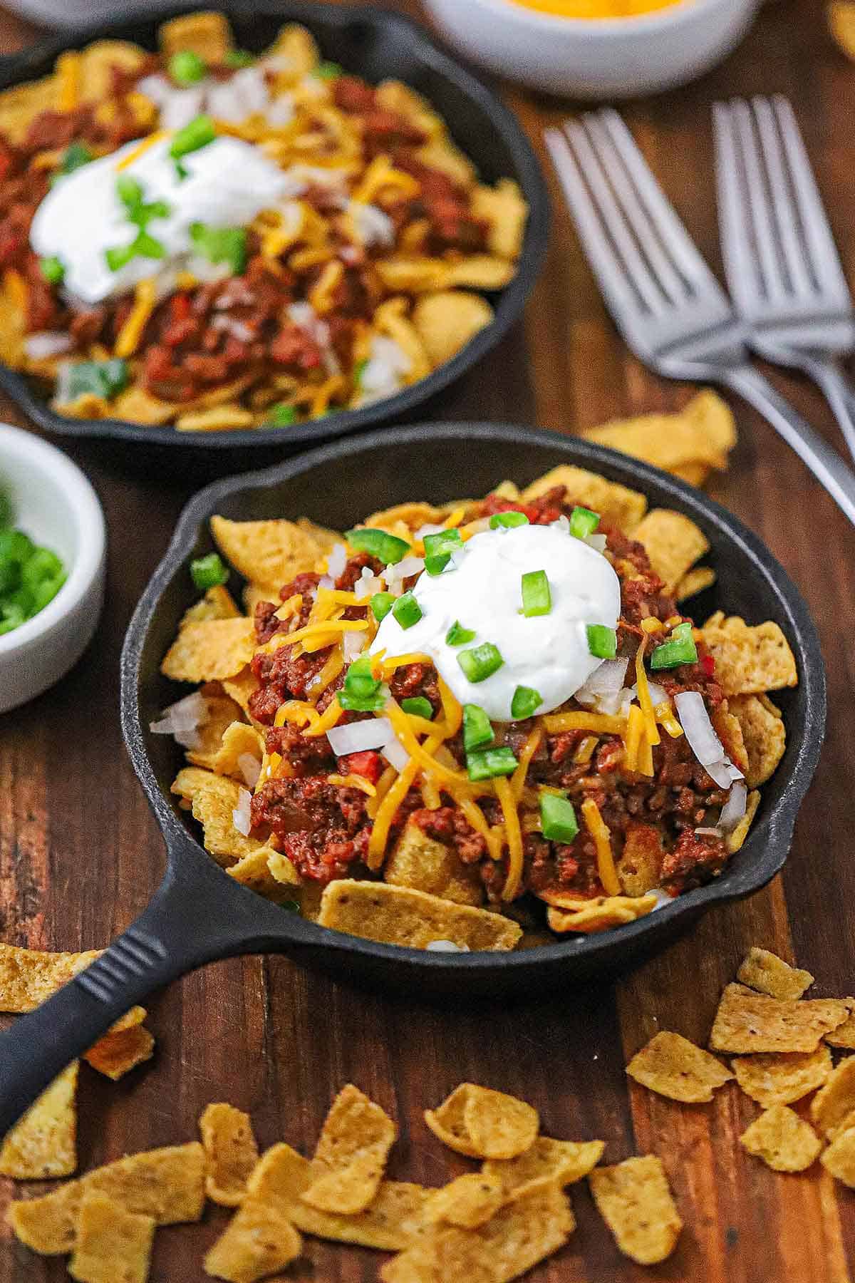 A straight-on view of two small cast-iron skillets both filled with individual servings of Classic Frito Pie topped with Beef Chili, shredded cheese, sour cream, and chopped jalapeños.