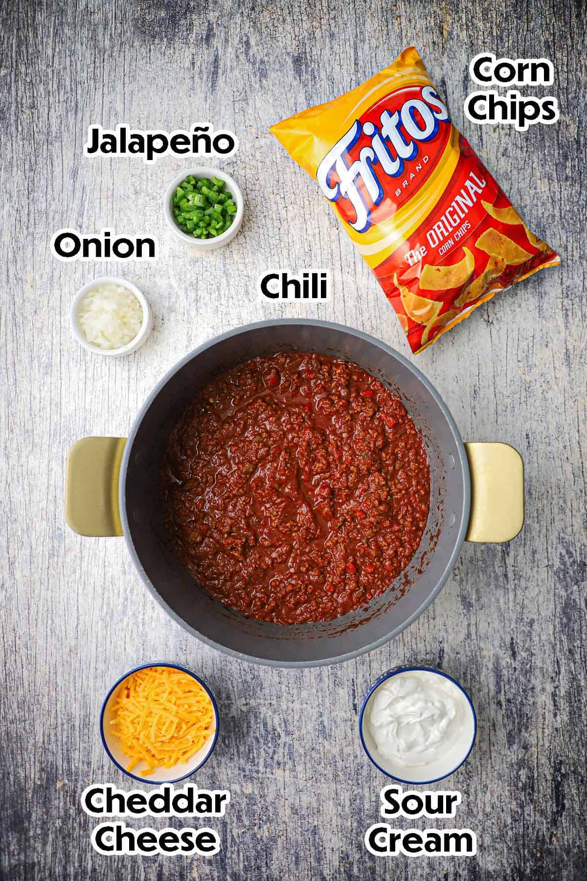 An arrangement of ingredients for Classic Frito Pie on a grey wooden background including a pot full of beef chili, a bag of Fritos, and small bowls of sour cream, cheddar cheese, onion, and jalapeños.