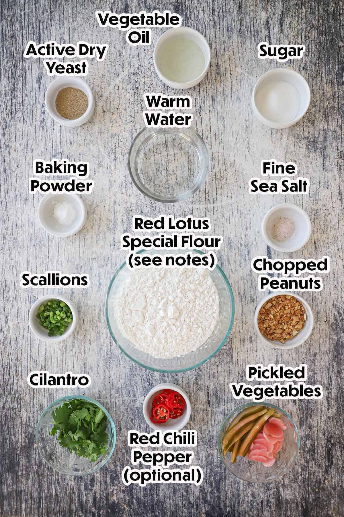 An arrangement of ingredients for homemade bao buns bread along with garnishes including flour, sugar, yeast, baking powder, oil, water, salt, scallions, peanuts, cilantro, pickled veggies, and peppers.