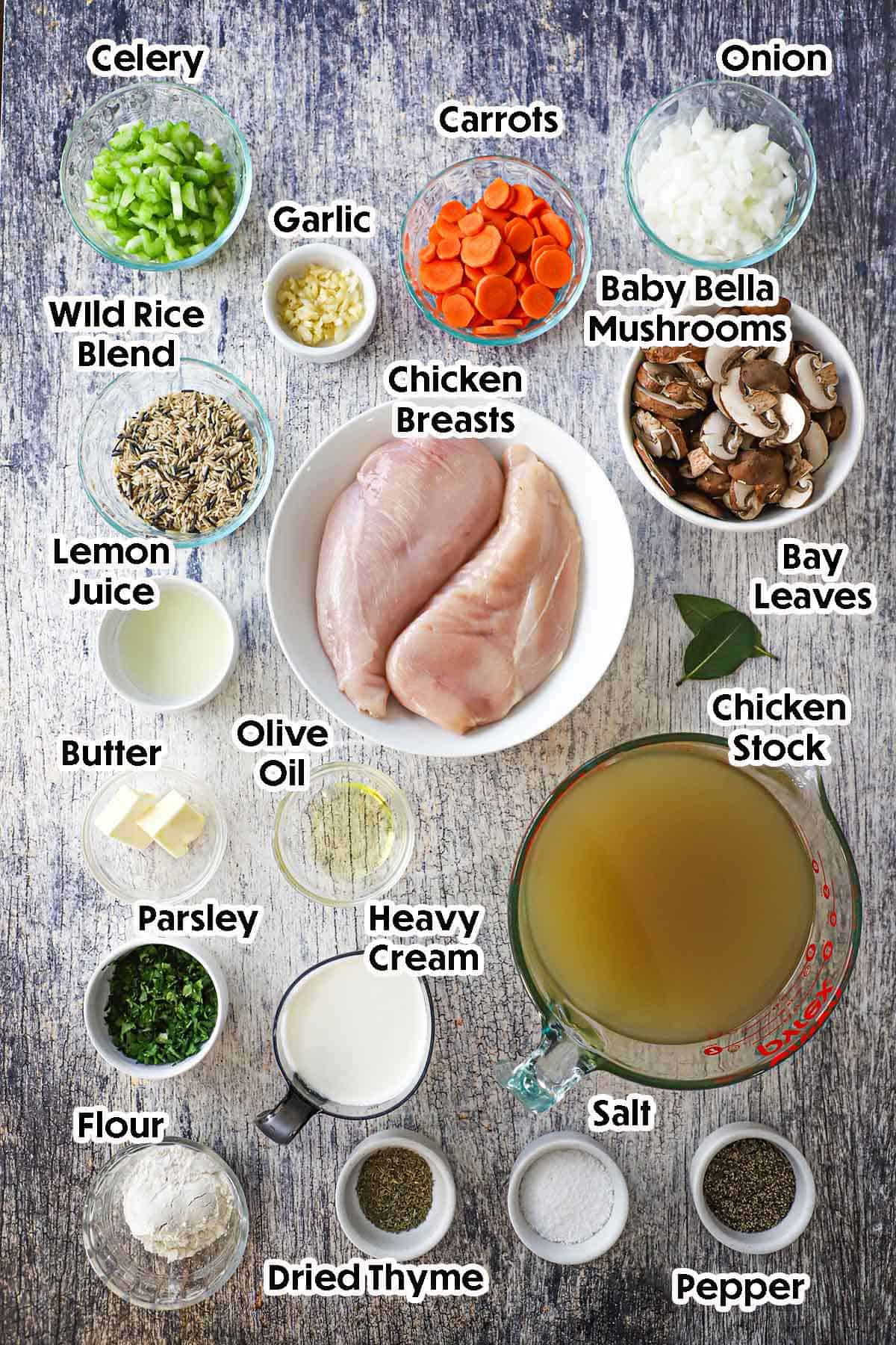 An overhead view of the ingredients for Creamy Chicken and Wild Rice Soup on a grey wooden background including chicken breasts, celery, carrots, onions, mushrooms, wild rice, chicken stock, cream, flour, butter, herbs, and seasonings.