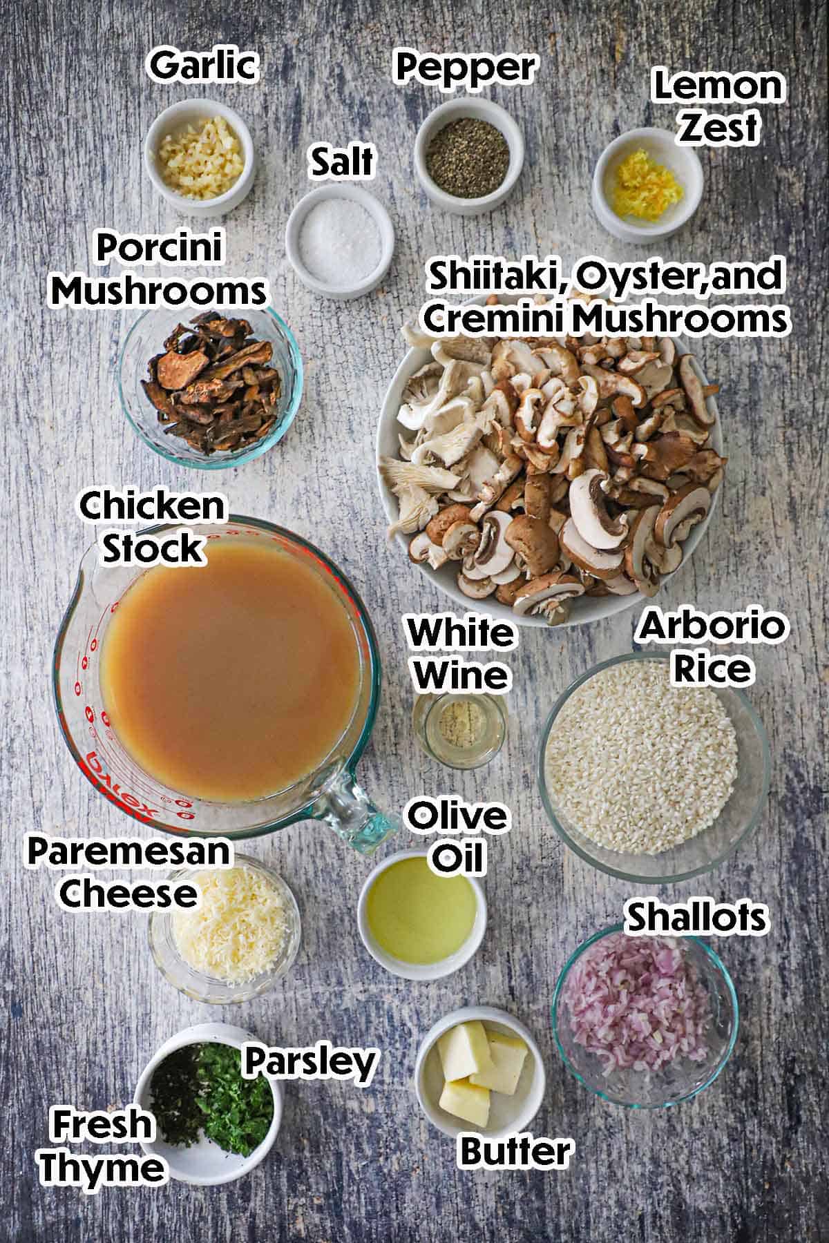 An arrangement of ingredients for wild mushroom risotto on a grey wooden background including sliced wild mushrooms, arborio rice, chicken stock, wine, shallots, Parmesan, oil, butter, garlic, herbs, and seasonings.