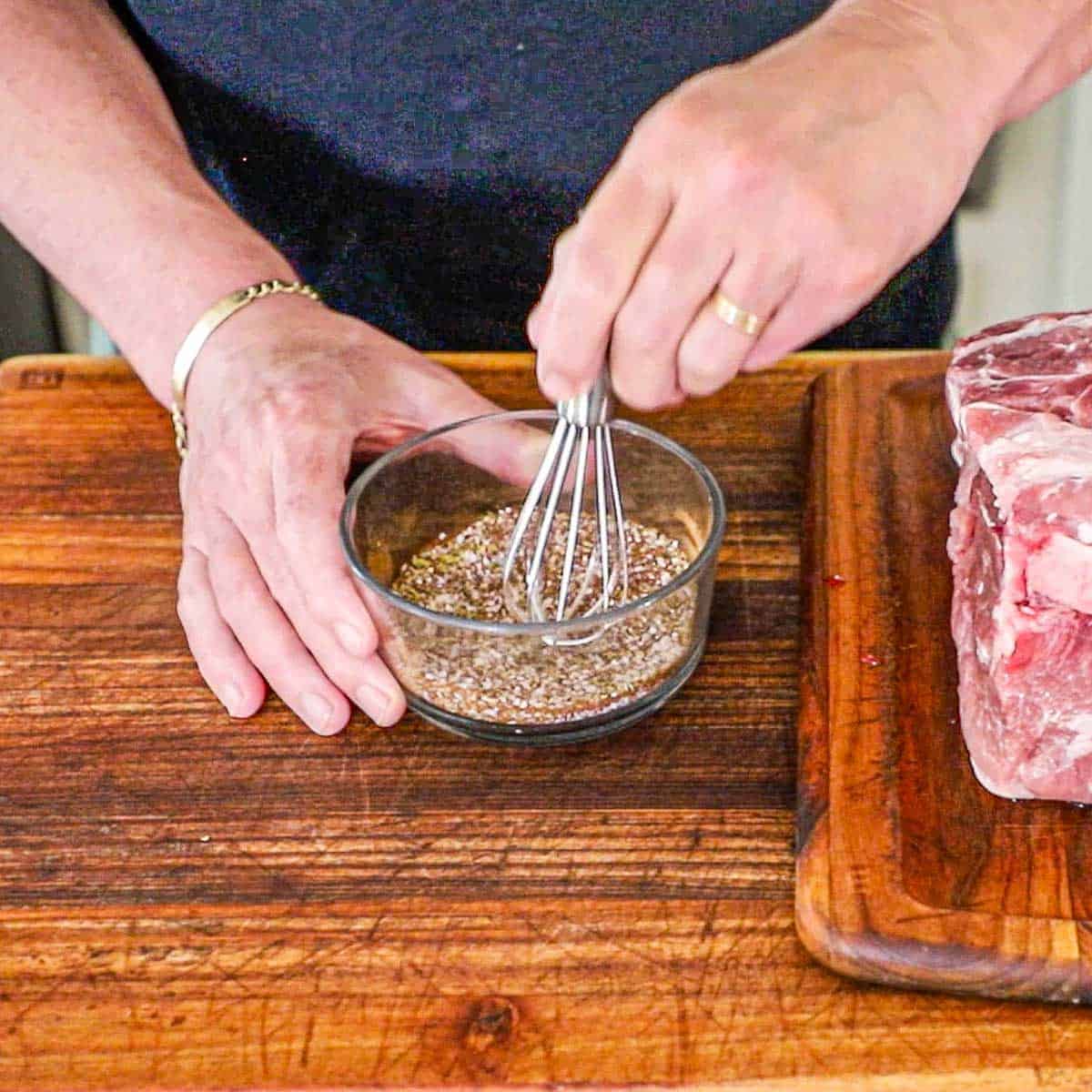 A person using a small whisk to combine salt with oregano, dried cumin, chili powder, and cayenne pepper in a small glass bowl next to an uncooked pork shoulder on a cutting board.