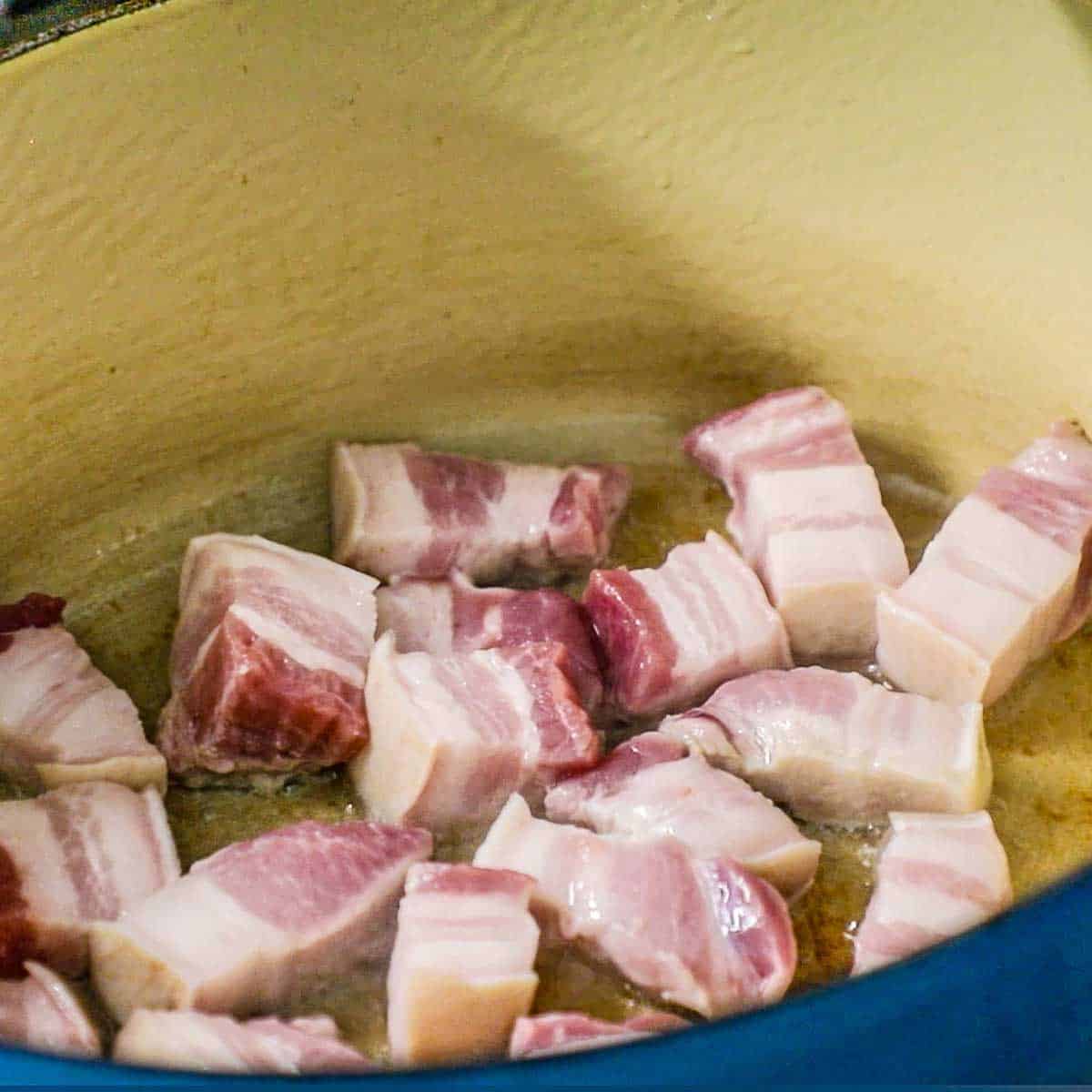 A close-up view of chunks of pork belly being seared in a large Dutch oven in a vegetable oil.