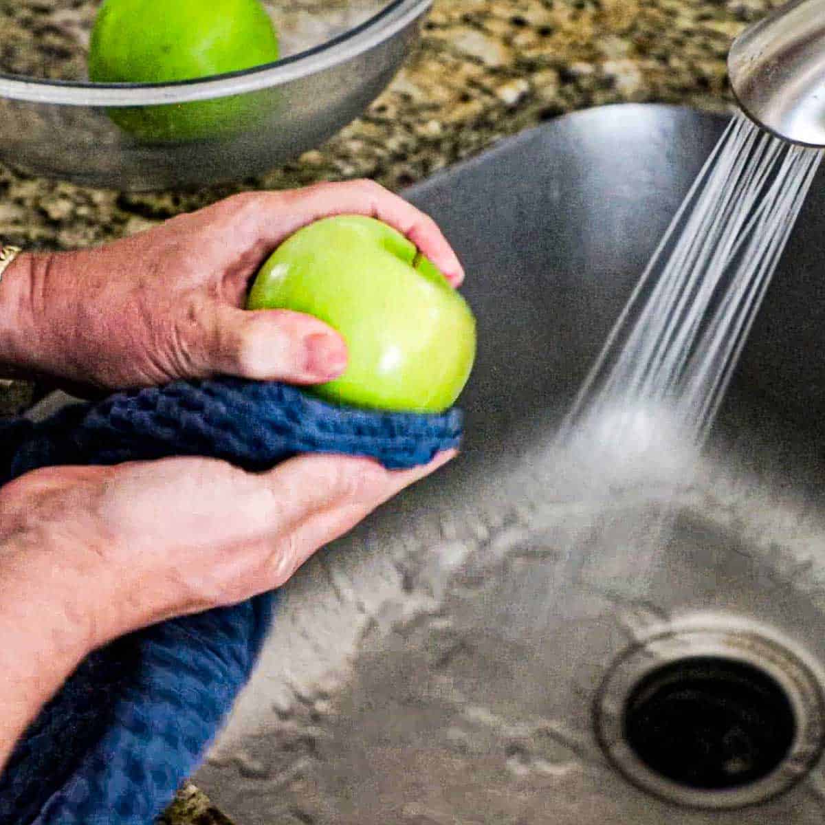 A person using a blue kitchen towel to scrub the wax from a Granny Smith apple that has been washed in hot tap water in the kitchen sink.