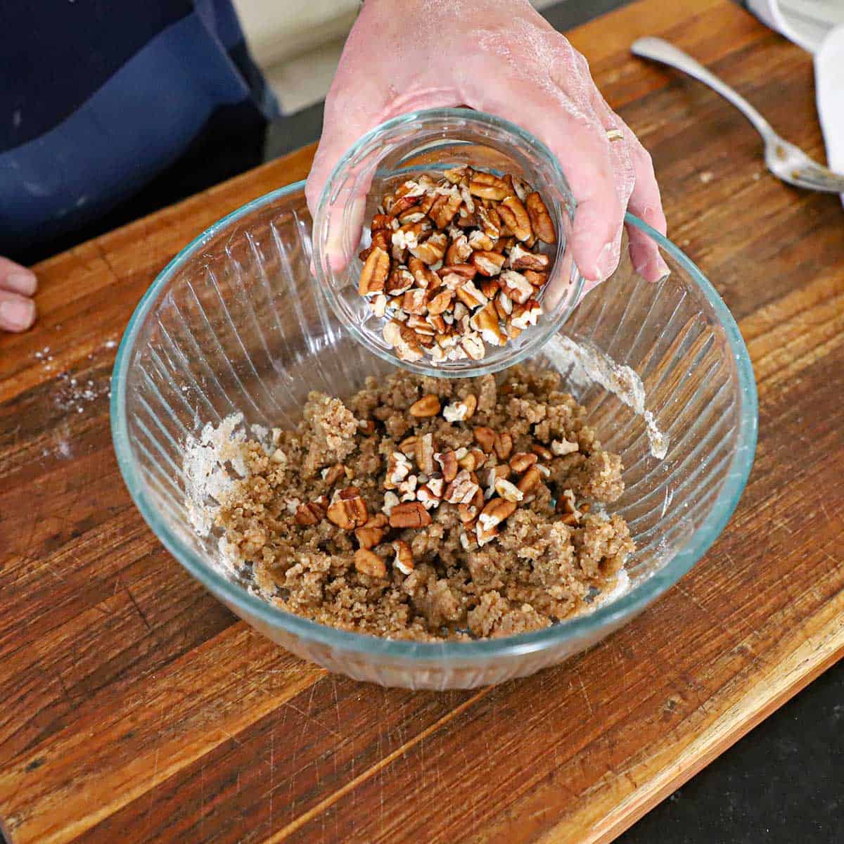 A person transferring chopped pecans into a mixture of melted butter, flour, and cinnamon in a glass bowl on a wooden cutting board.