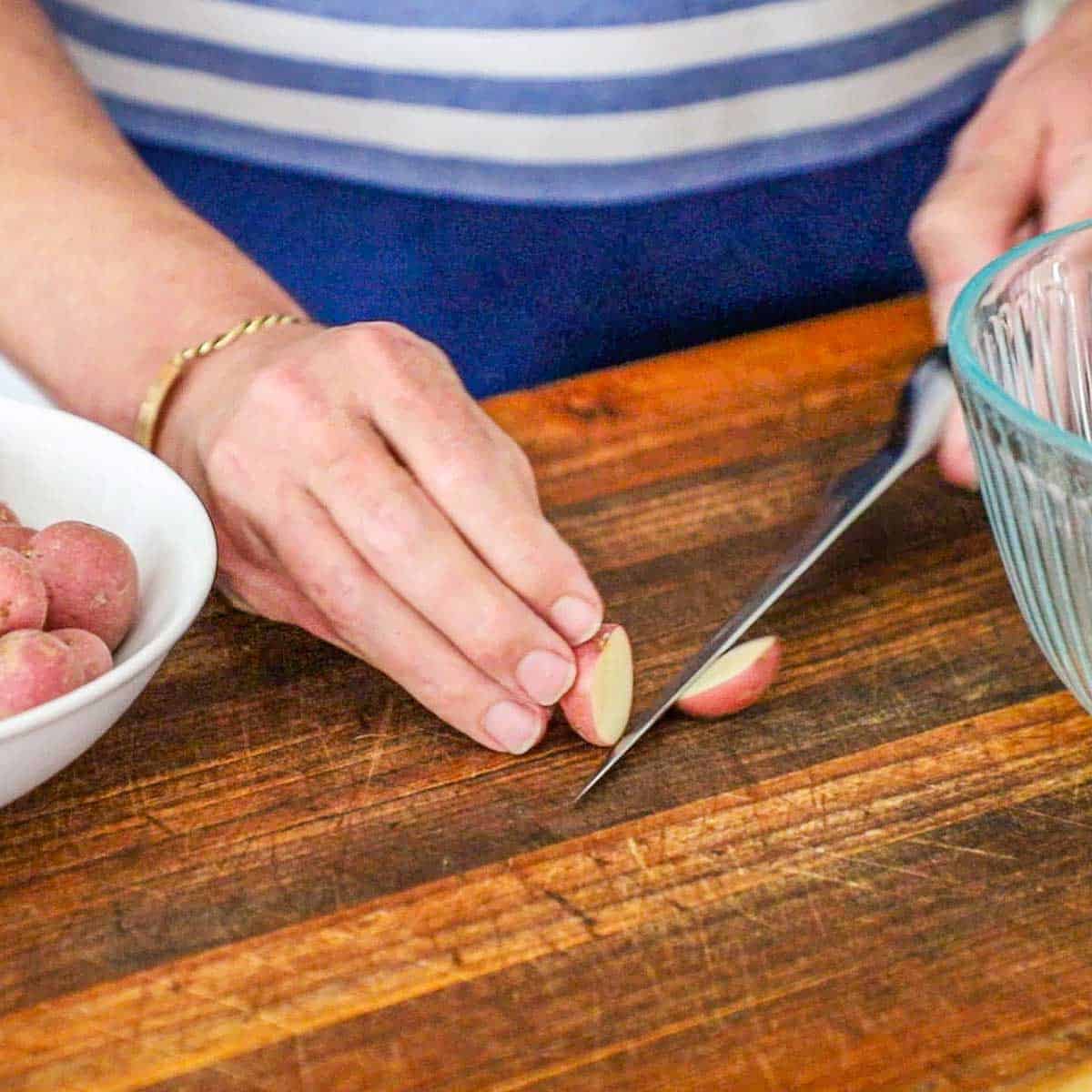 A person using a knife to cut a baby red potato in half on a large wooden cutting board with a glass bowl sitting nearby.