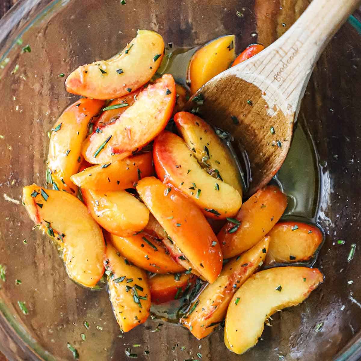 An overhead view of sliced peaches that have been tossed in a fresh herb marinade in a glass bowl.