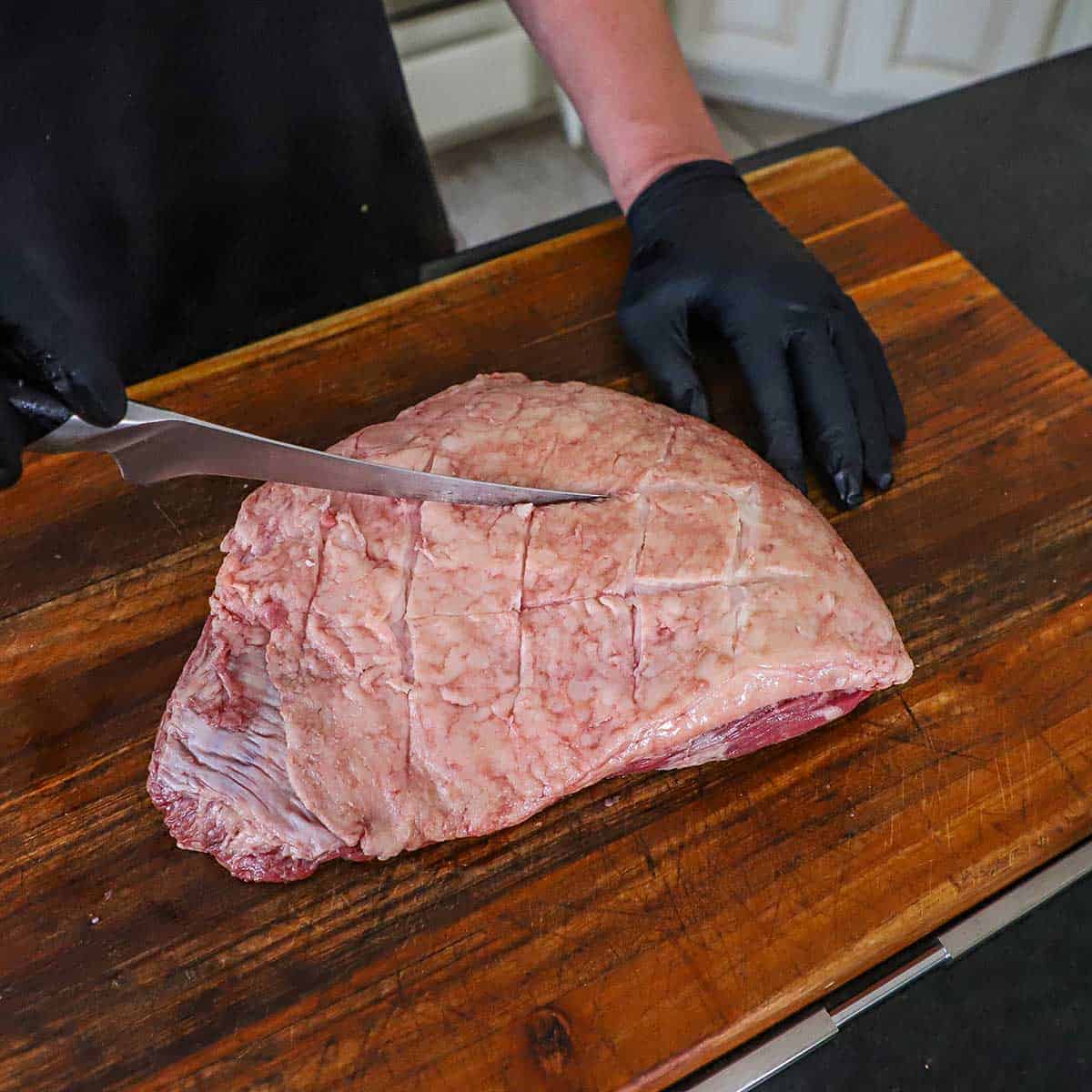 A person using a sharp knife to score a diamond shape into the top fat cap of an uncooked picanha steak resting on a cutting board.