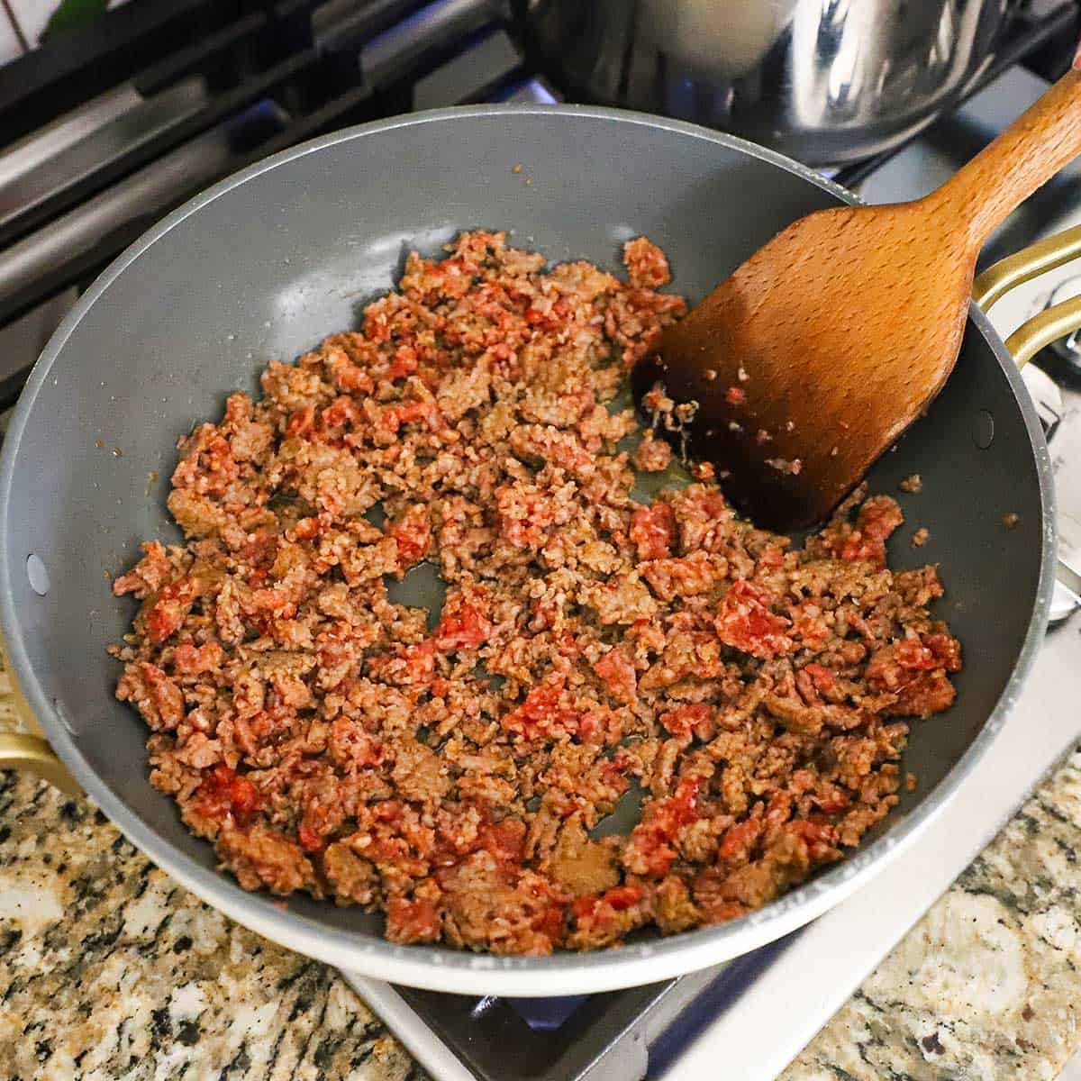 A person using a wooden spatula to stir crumbled Italian sausage that is being cooked in a large skillet on a gas stove.