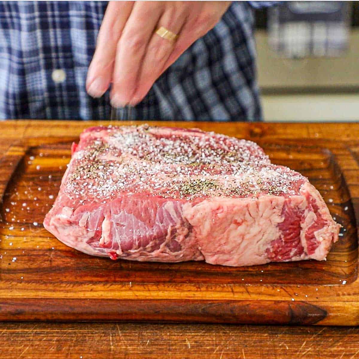 A person using his fingers to sprinkle salt and pepper over the surface of an uncooked beef chuck roast that is resting on a wooden cutting board.
