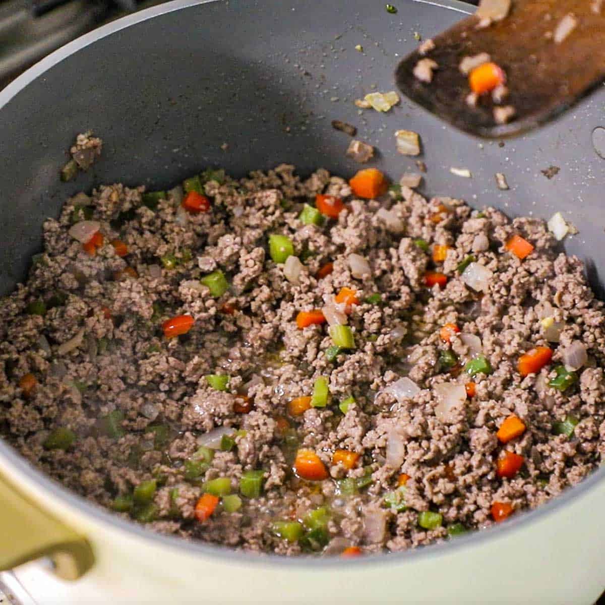 A close-up view of a pot that is filled with cooked ground beef combined with sautéed onion, bell peppers, and chopped jalapeños all in a large pot on a gas stove.