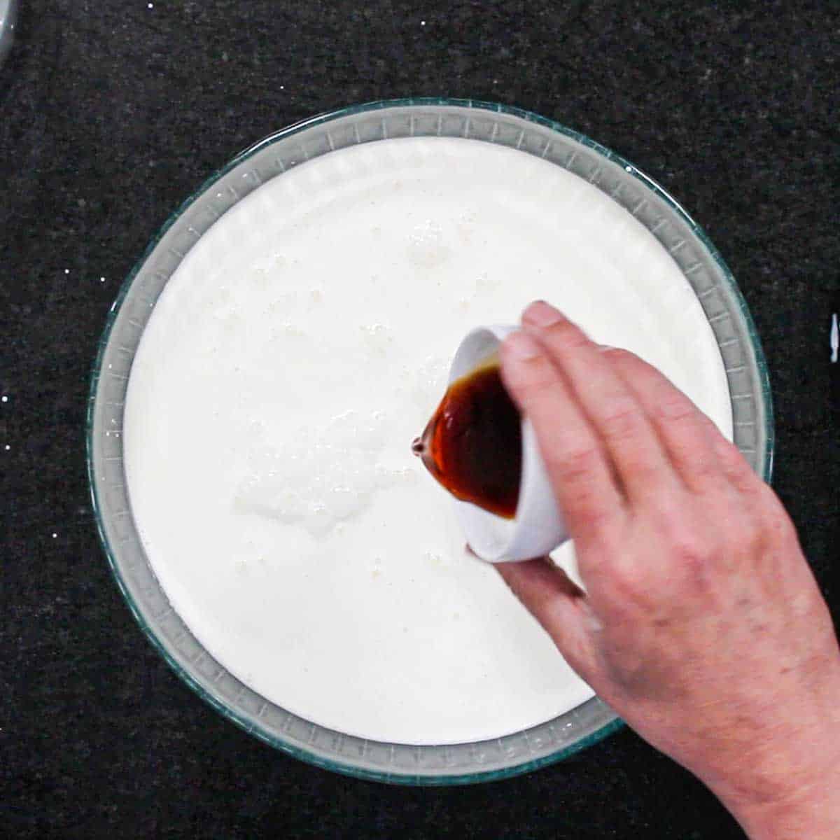 A person pouring vanilla extract from a small white bowl into a large glass bowl filled with cream combined with milk and sugar.