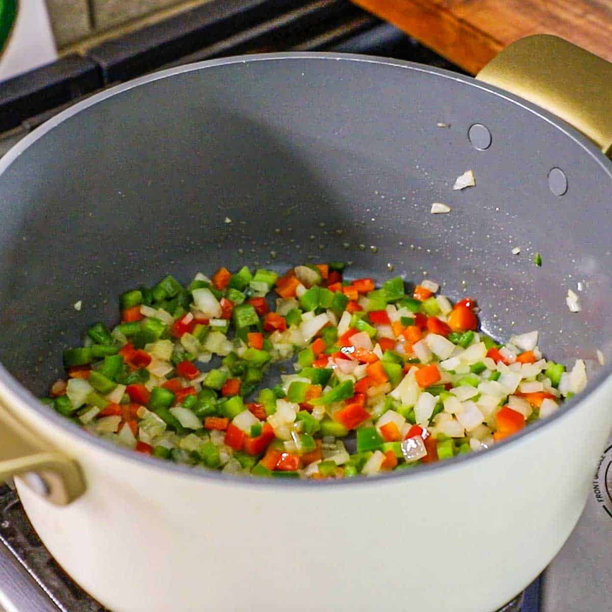 A large pot filled with chopped onion, red bell pepper, green bell pepper, and jalapeño being sautéed in olive oil on a gas stove.