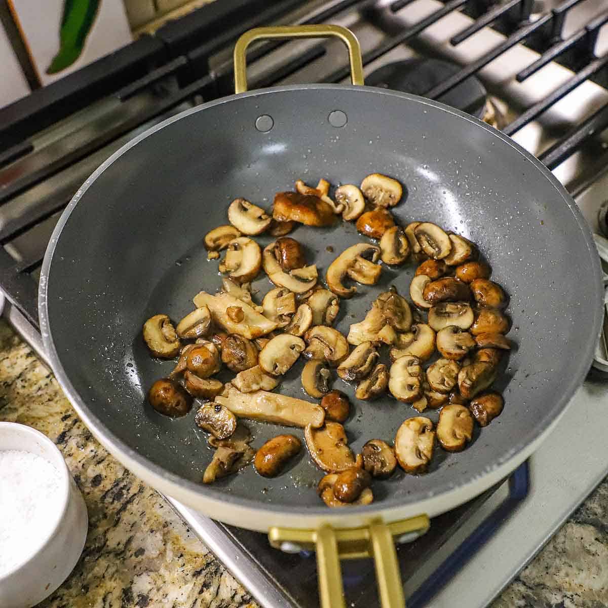 Sliced wild mushrooms being saut&eacute;ed in a large non-stick skillet on a gas stove.