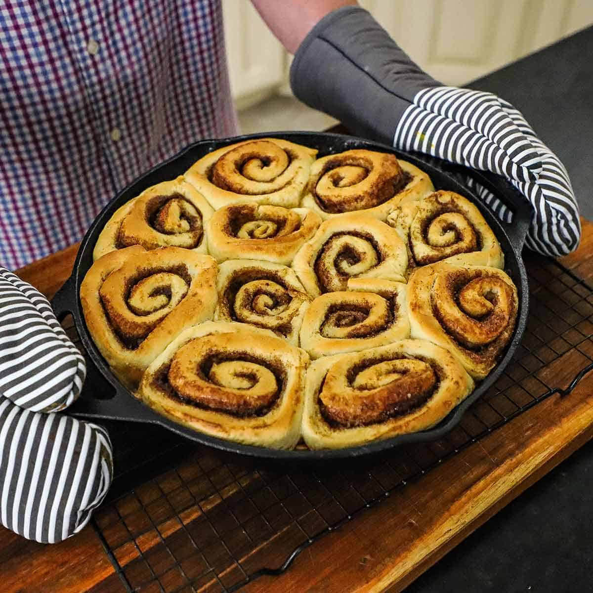 A person wearing oven mitts holding the handles of a large cast-iron skillet that is filled with freshly baked cinnamon rolls and is resting on a baking rack on a wooden cutting board.