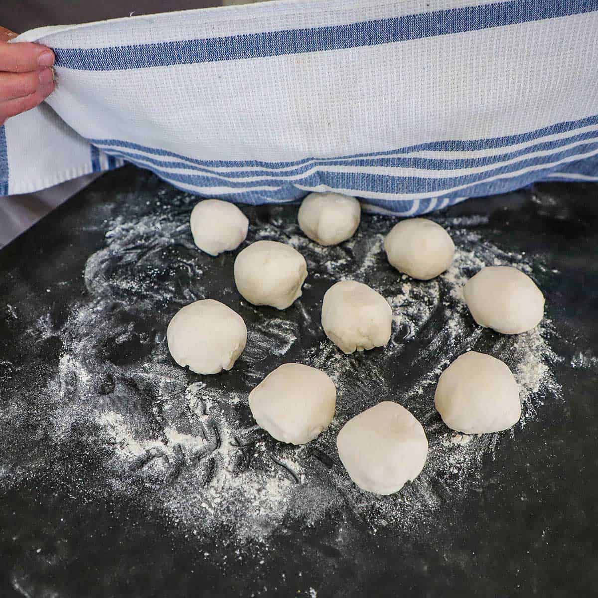 A person holding a kitchen towel over ten balls of bao dough on a lightly floured counter.