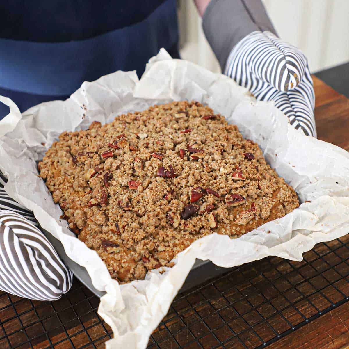 A person wearing oven mitts holding a square baking pan that is lined with parchment paper and filled with a freshly baked pumpkin spice coffee cake.