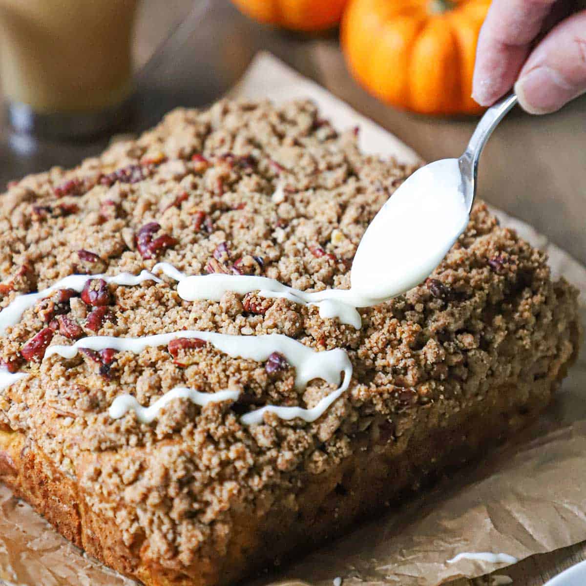 A person using a spoon to drizzle maple glaze over the top of a square pumpkin spice coffee cake topped with a pecan streusel.