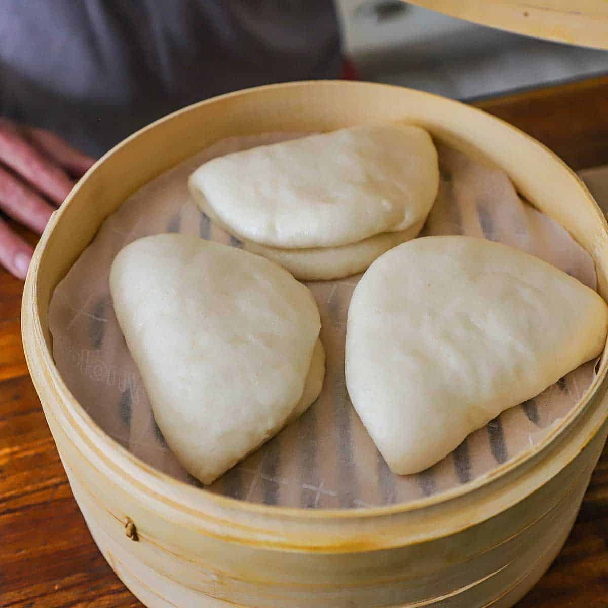 A person lifting a the lid off of a bamboo steamer revealing three freshly steamed bao buns resting on parchment paper.