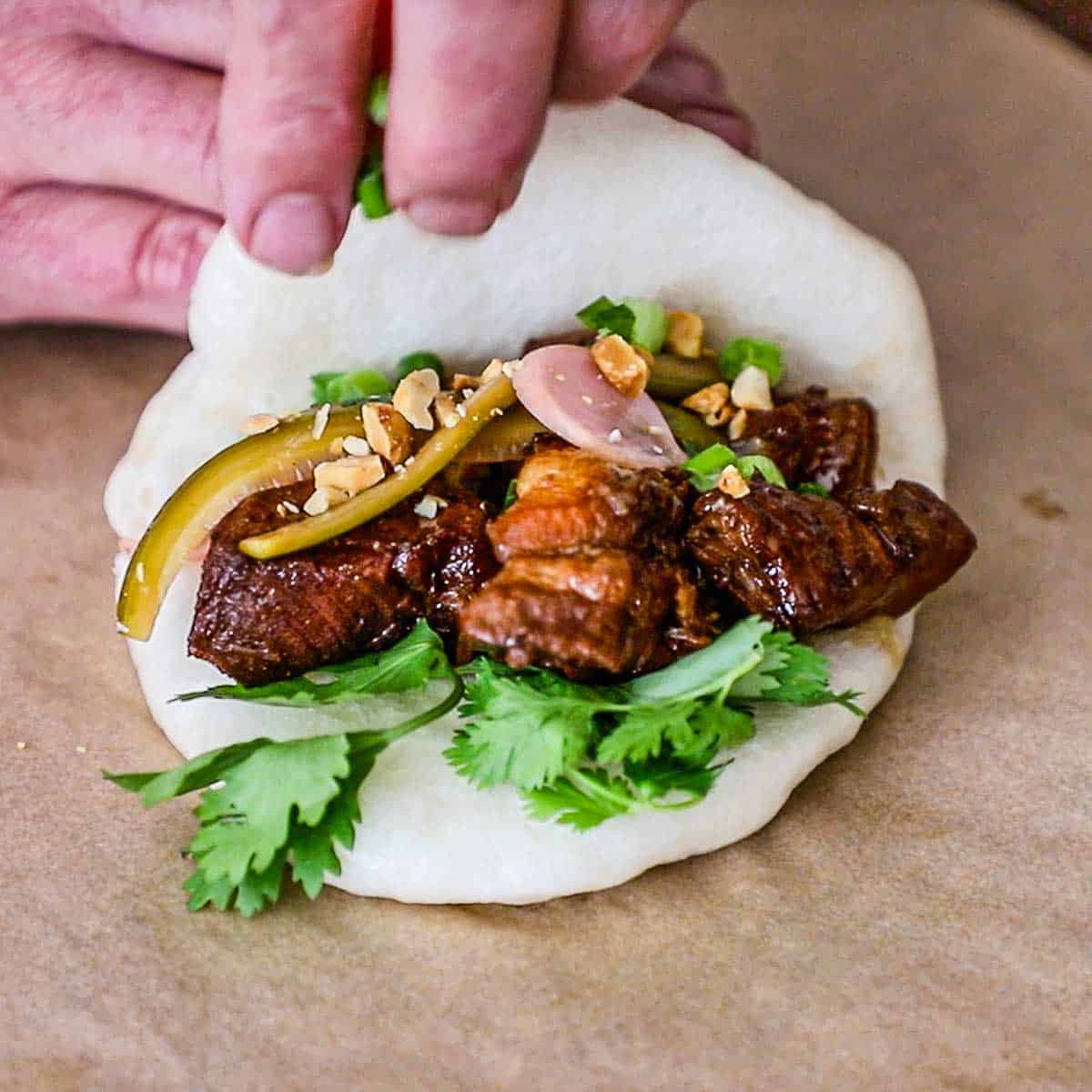 A person sprinkling chopped scallions over the top of braised pork belly chunks resting inside a freshly steamed bao bun garnished with chopped cilantro, pickled vegetables, and chopped peanuts.