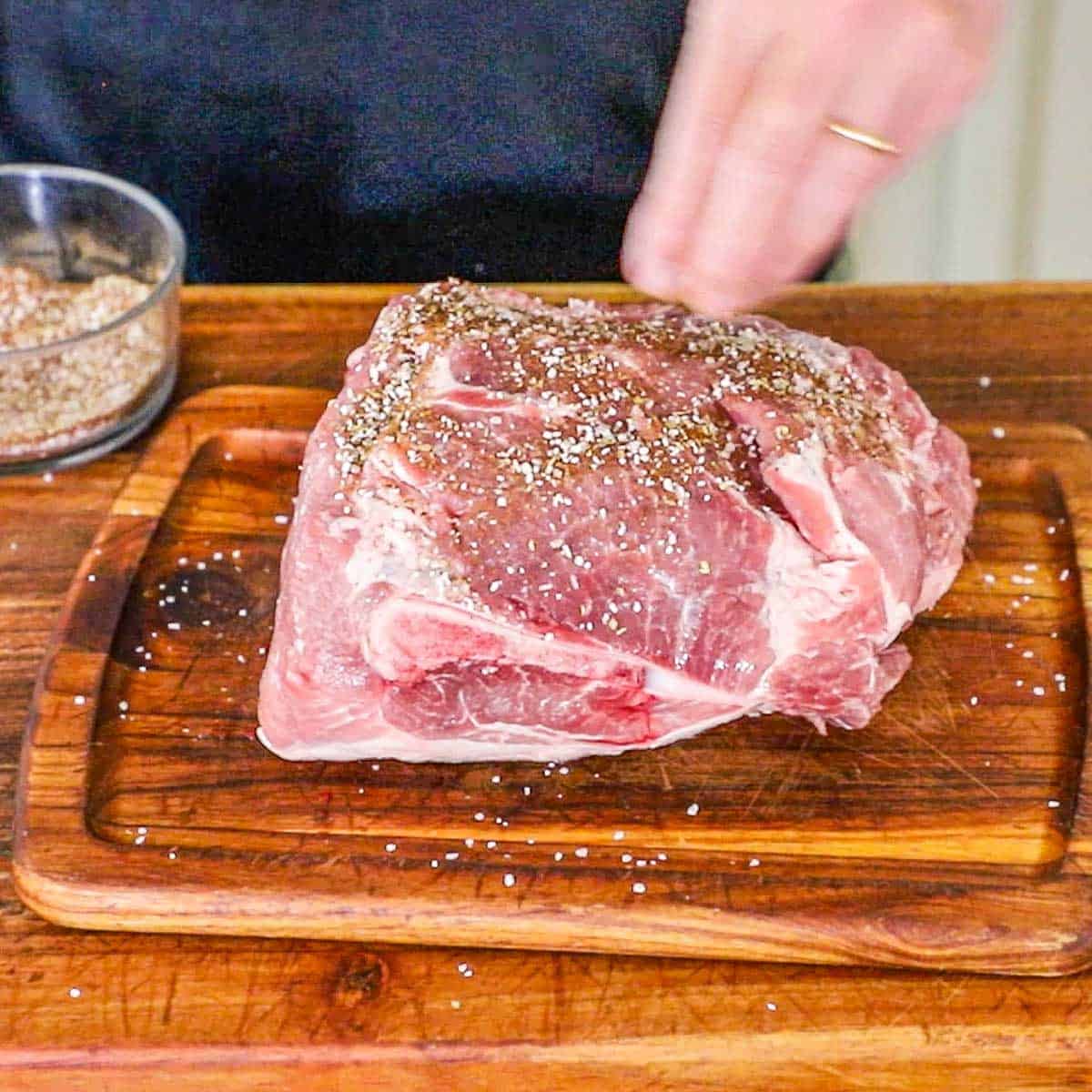 A person sprinkling an uncooked pork shoulder with a spice rub while it's resting on a wooden cutting board.