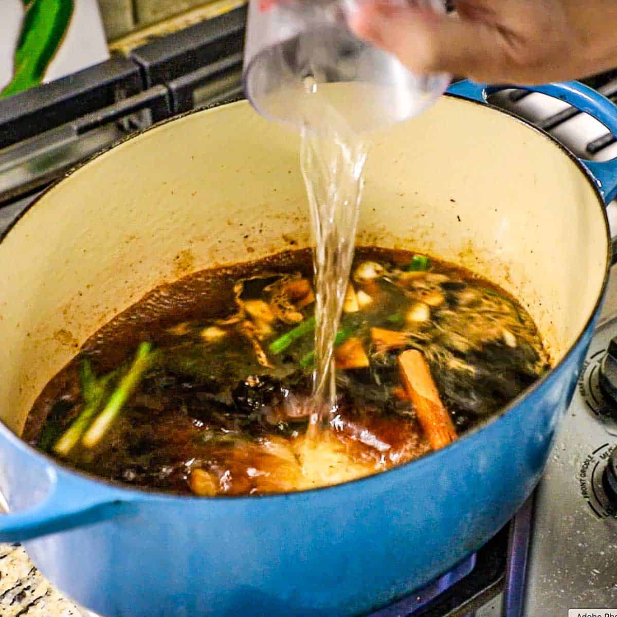 A person pouring water from a plastic measuring cup into a large Dutch oven filled with a simmering Asian soy and cinnamon braising liquid for pork belly.