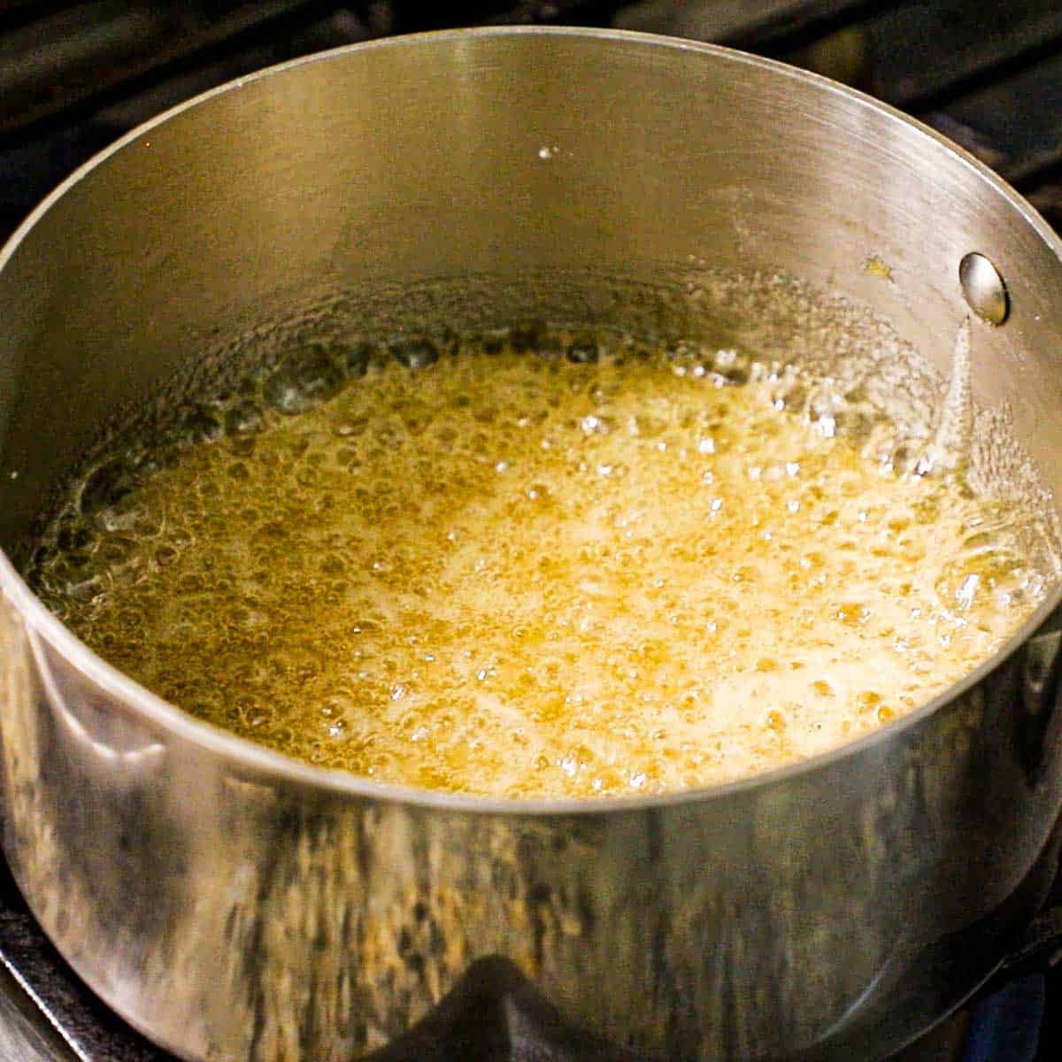 A close-up view of a silver saucepan that contains a gurgling mixture dissolved sugar and honey on a gas stove.