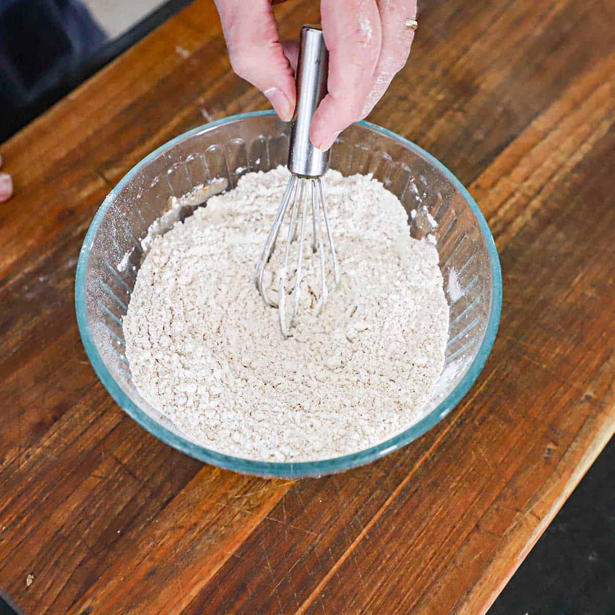 A person using a small whisk to combine flour with baking powder, baking soda, and spices in a glass bowl on a wooden cutting board.