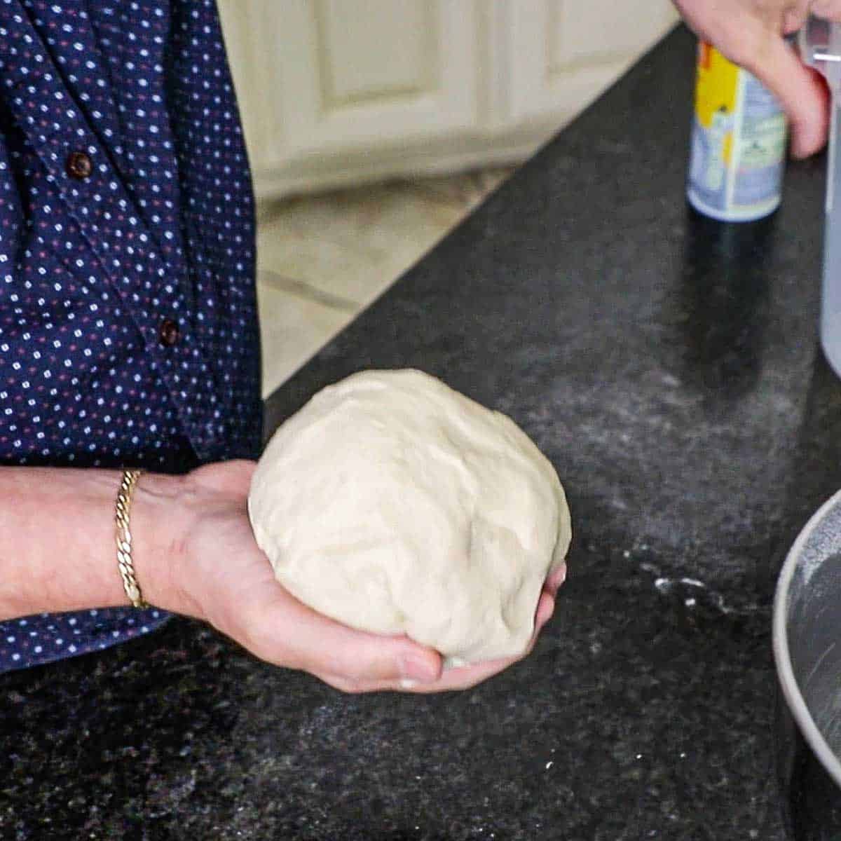 A person holding a ball of telera dough in his hand over a black marble countertop.