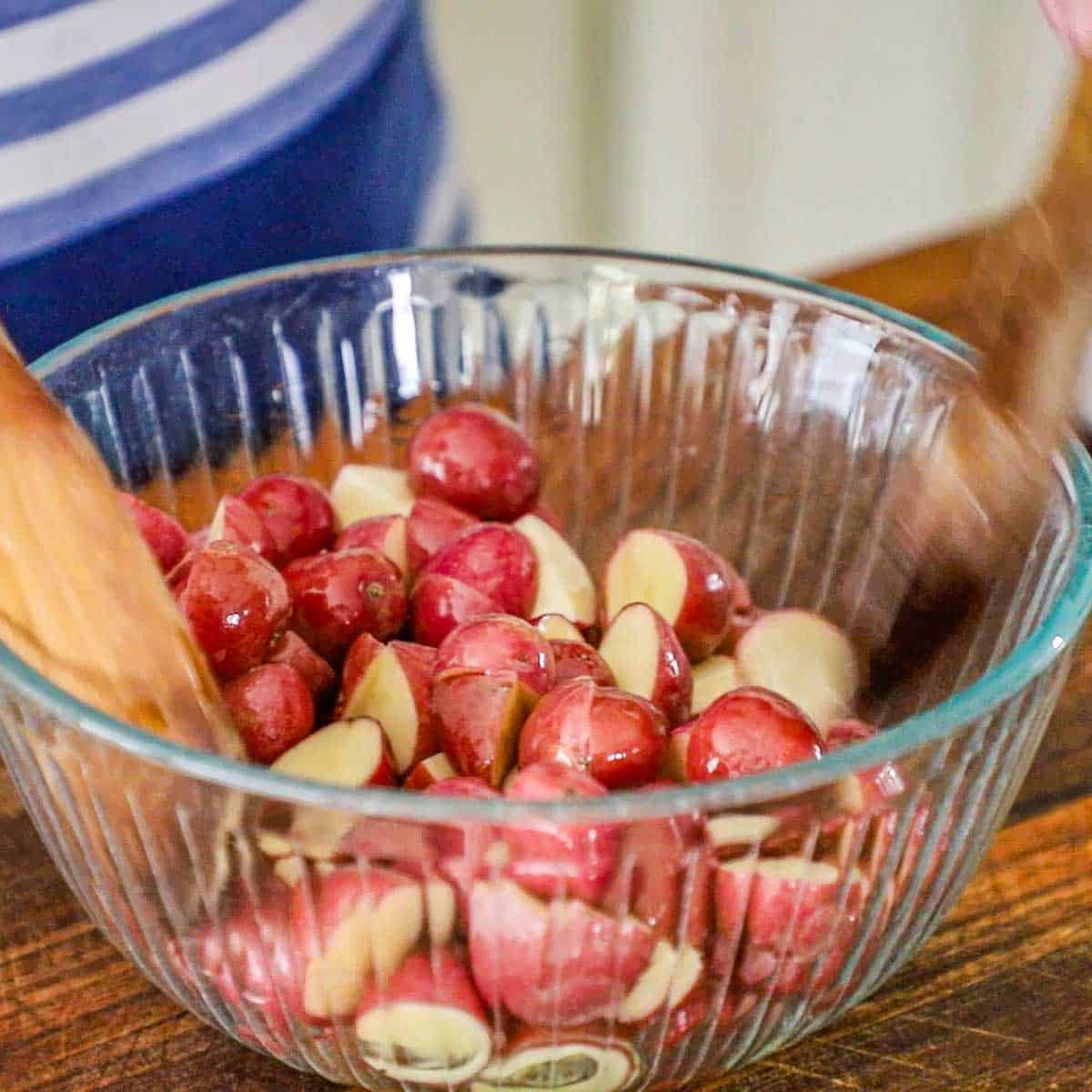 A person using two large wooden spoons to toss halved baby red potatoes with olive oil in a large glass bowl on a wooden cutting board.