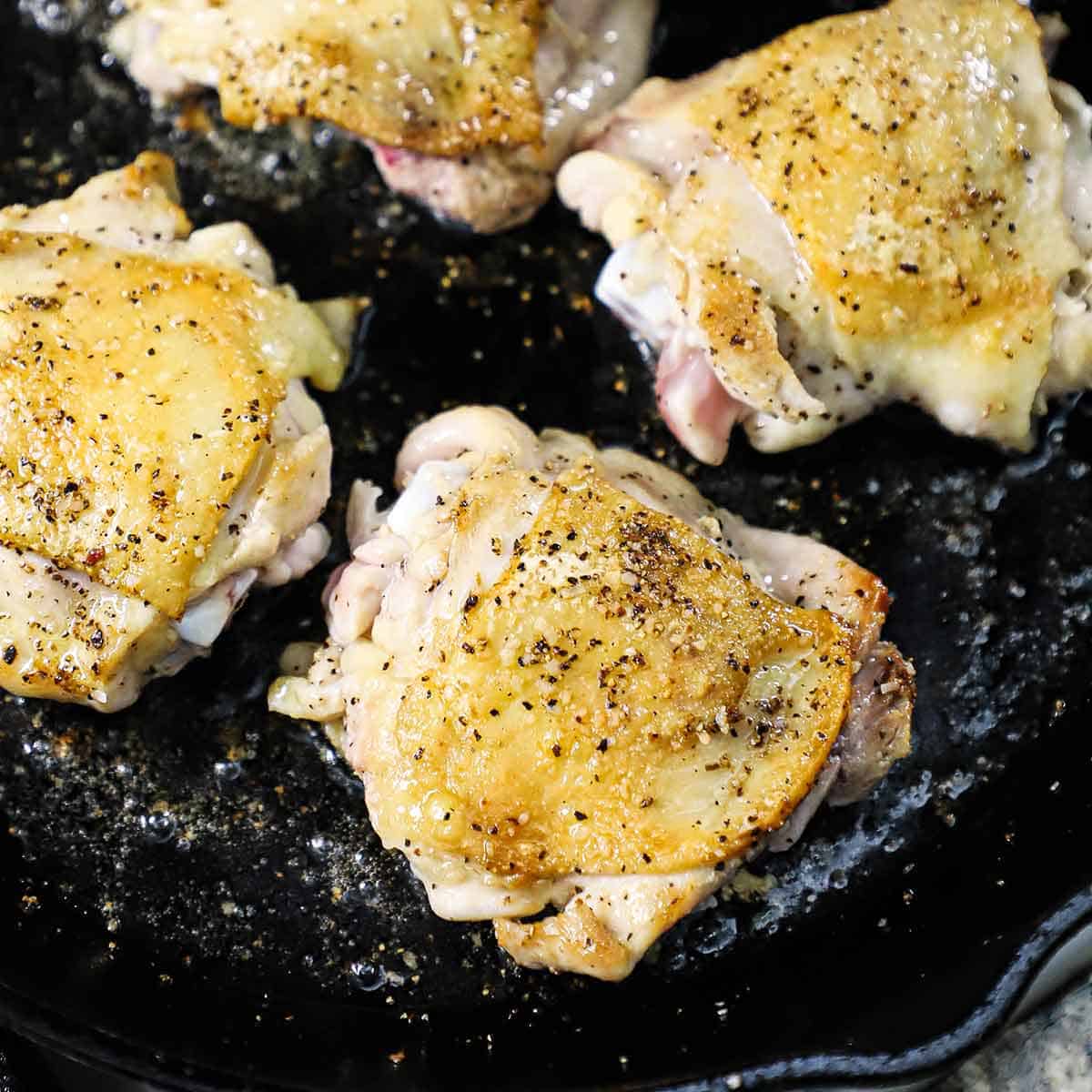 A close-up view of four skin-on chicken thighs that have been seasoned with salt and pepper and are being seared and crisped in a large cast-iron skillet with oil.