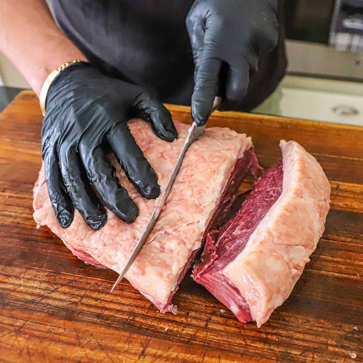 A person using a sharp butcher's knife to slice thick slabs from a whole picanha roast on a large cutting board.