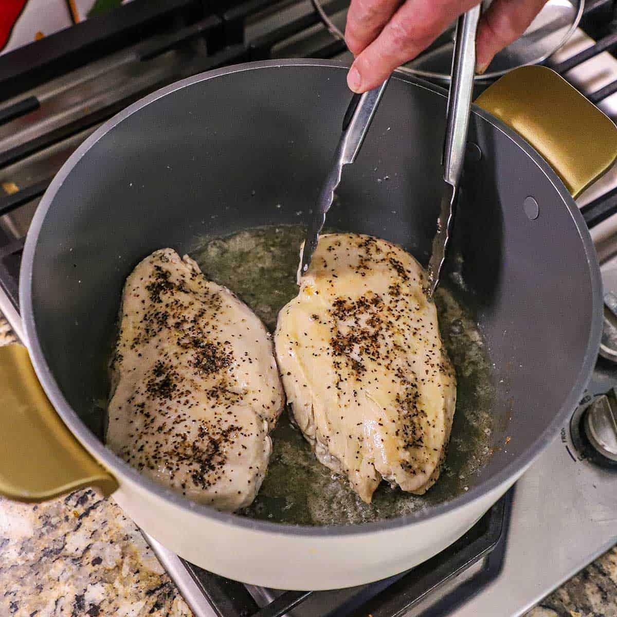 A person using a pair of metal tongs to turn two seasoned chicken breasts that are being browned in a large pot in olive oil on a gas stove.