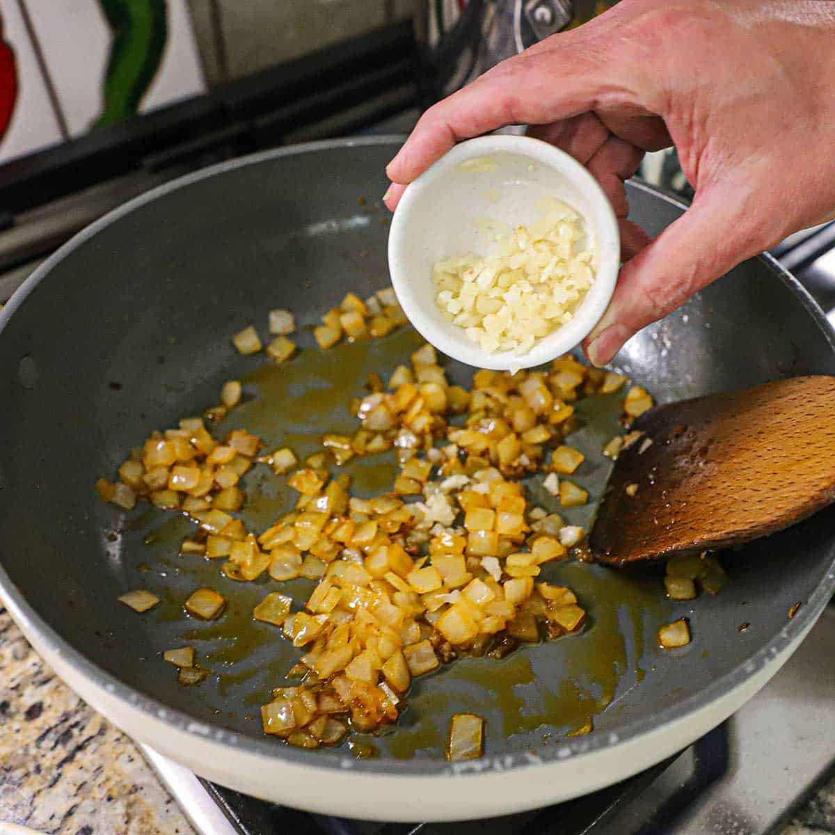 A person dumping minced garlic from a small white bowl into a large skillet that has chopped onion that has been sautéed in oil on a gas stove.