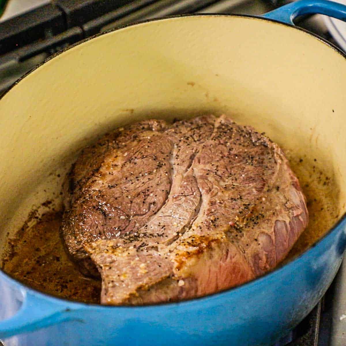 A seasoned beef chuck roast being seared in a large oval Dutch oven on a gas stove.
