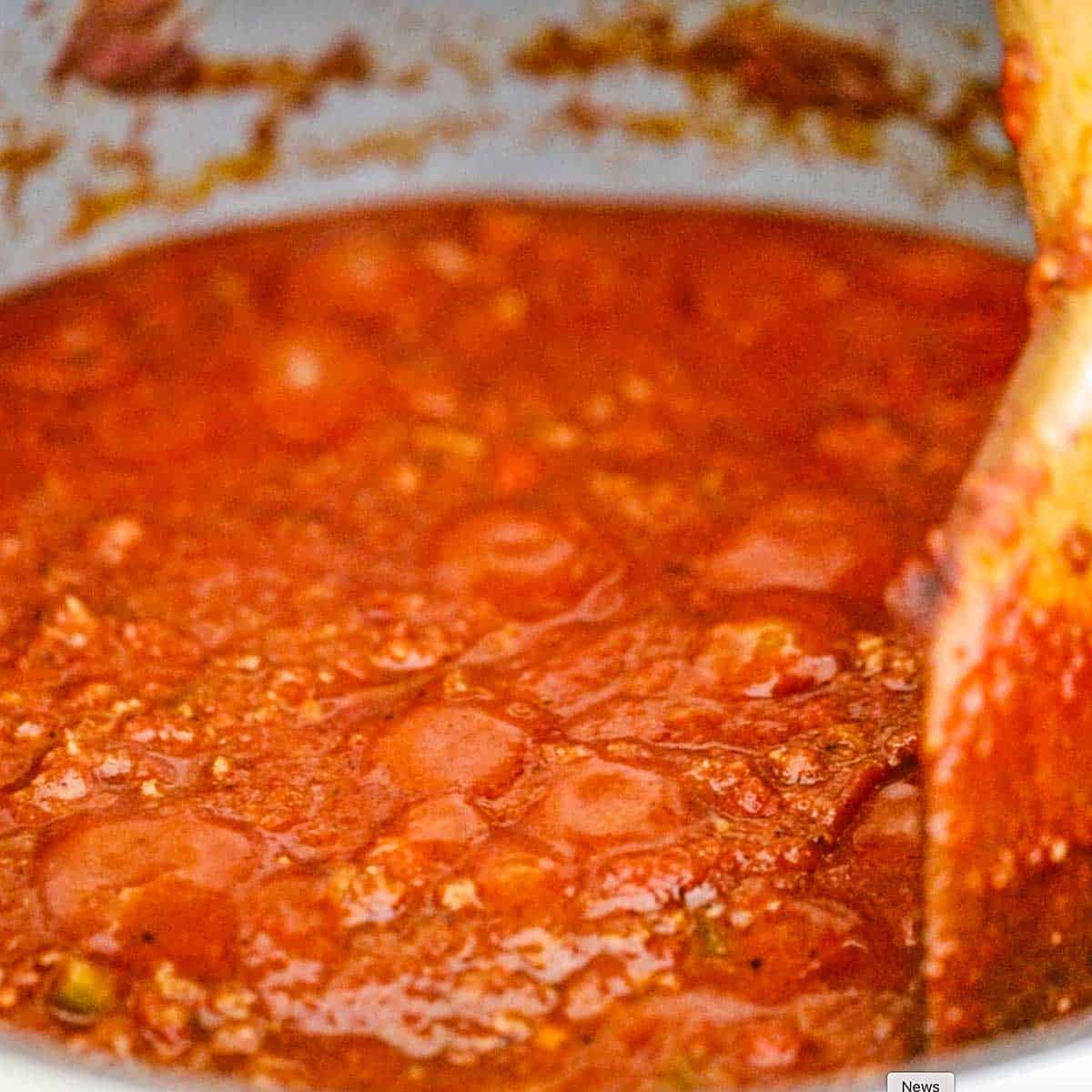 A close-up view of a pot of simmering beef chili with a wooden spatula being used to stir it.