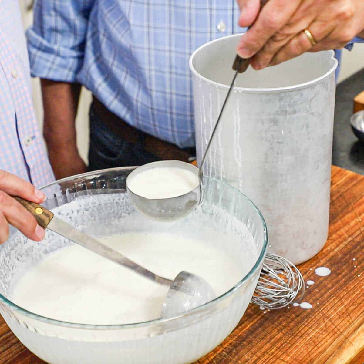 Two people both using a ladle to transfer a vanilla cream from a large glass bowl into a large metal ice cream maker container.