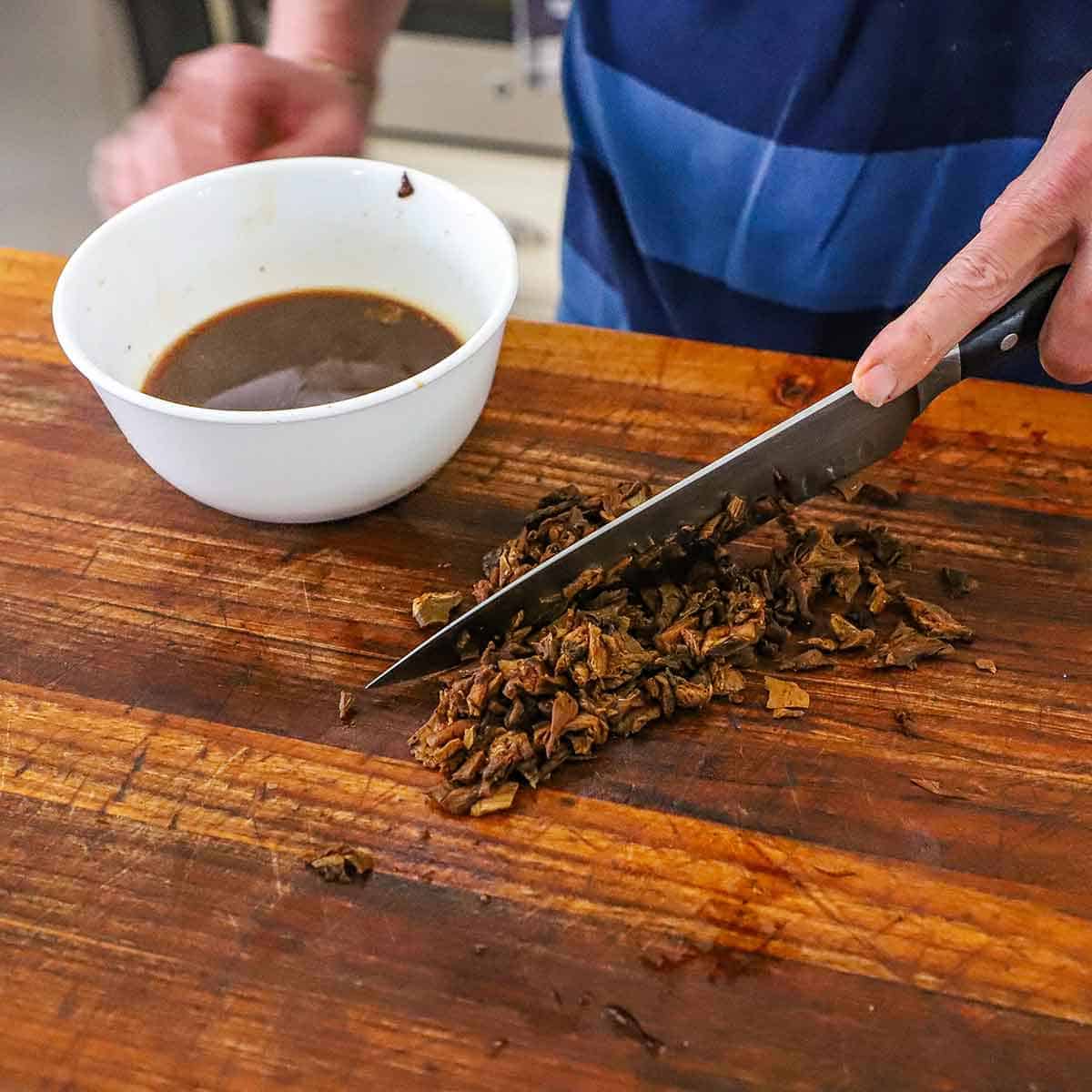 A person using a chef's knife to chop reconstituted dried porcini mushrooms on a large wooden cutting board with a white bowl nearby containing the soaking liquid.