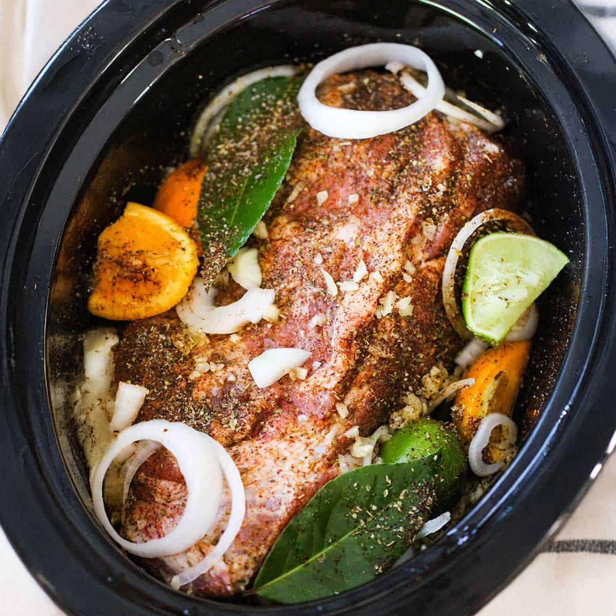 An overhead view of a large slow cooker filled with a seasoned, uncooked pork should, along with squeezed citrus, slices of onions, minced garic, and bay leaves