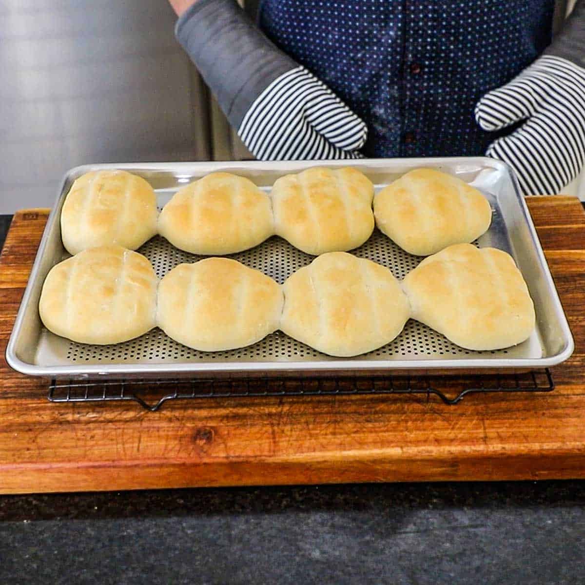 A person wearing oven mitts standing behind a kitchen counter with a baking pan filled with homemade telera rolls on a baking rack resting on a wooden cutting board.