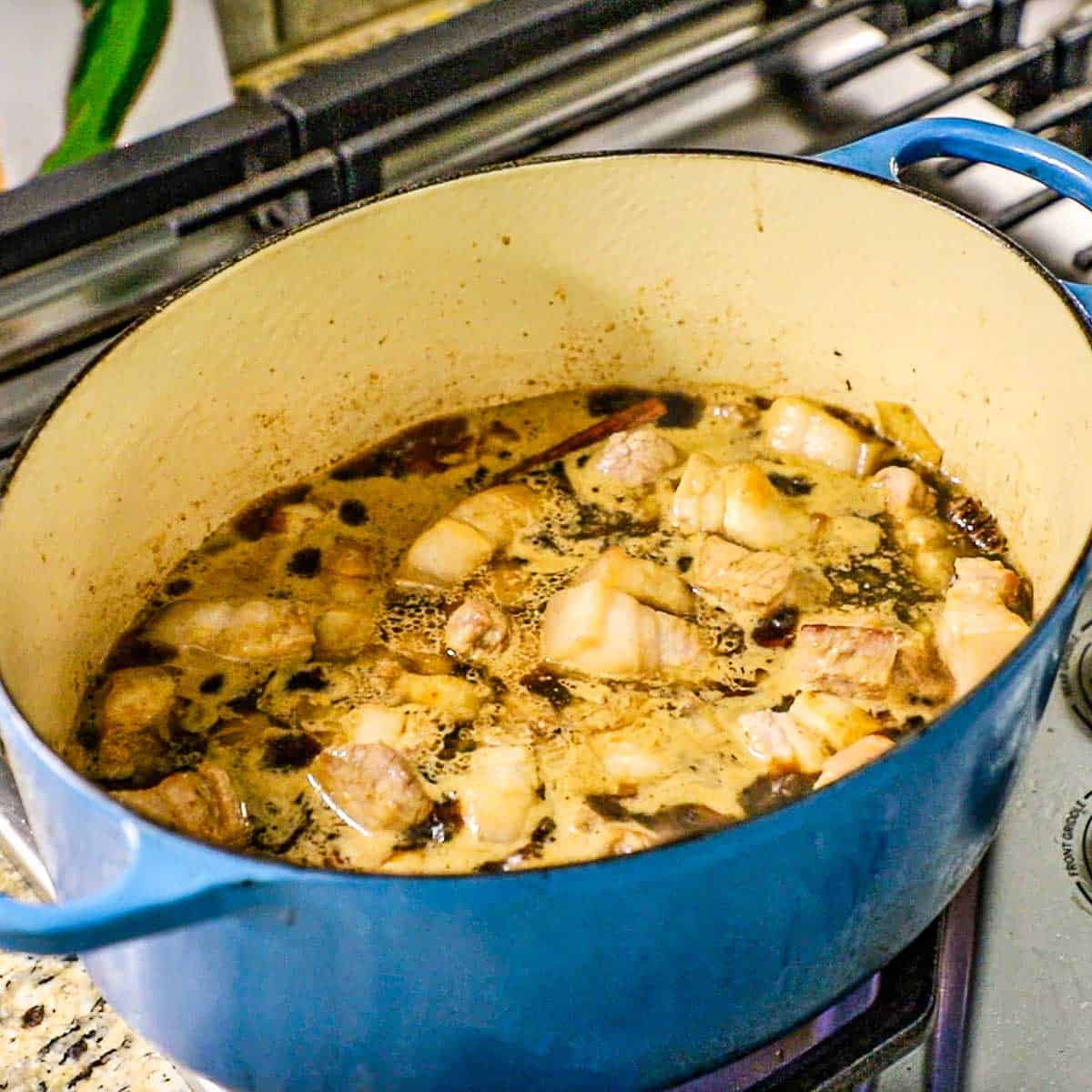 A straight-on view of a large Dutch oven filled with a simmering dark sauce with chunks of seared pork belly in it.