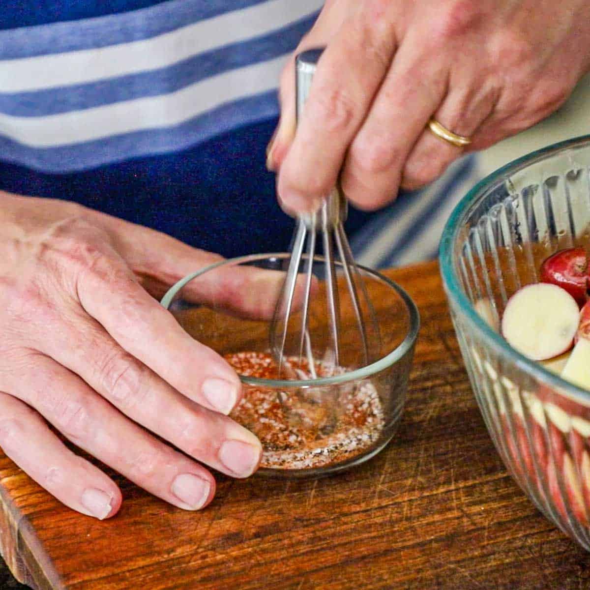 A person using a small whisk to combine paprika, garlic powder, salt, and pepper in a small glass bowl sitting next to a large glass bowl filled with oiled baby red potatoes that have been halved.