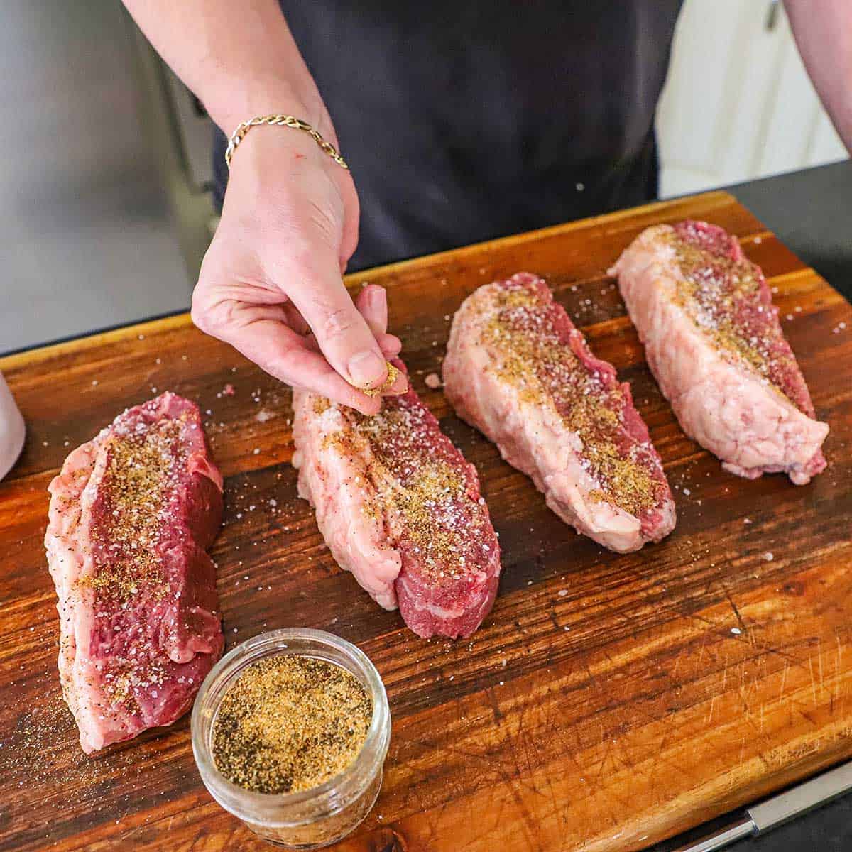 A person using his fingers to sprinkle beef rub over the surface of four large steaks of uncooked picanha on a wooden cutting board.
