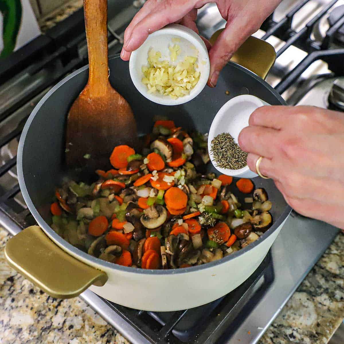 A person holding a small bowl of minced garlic in one hand and another small white bowl filled with dried thyme both over a large pot filled with sautéed carrots, celery, onion, and mushrooms.