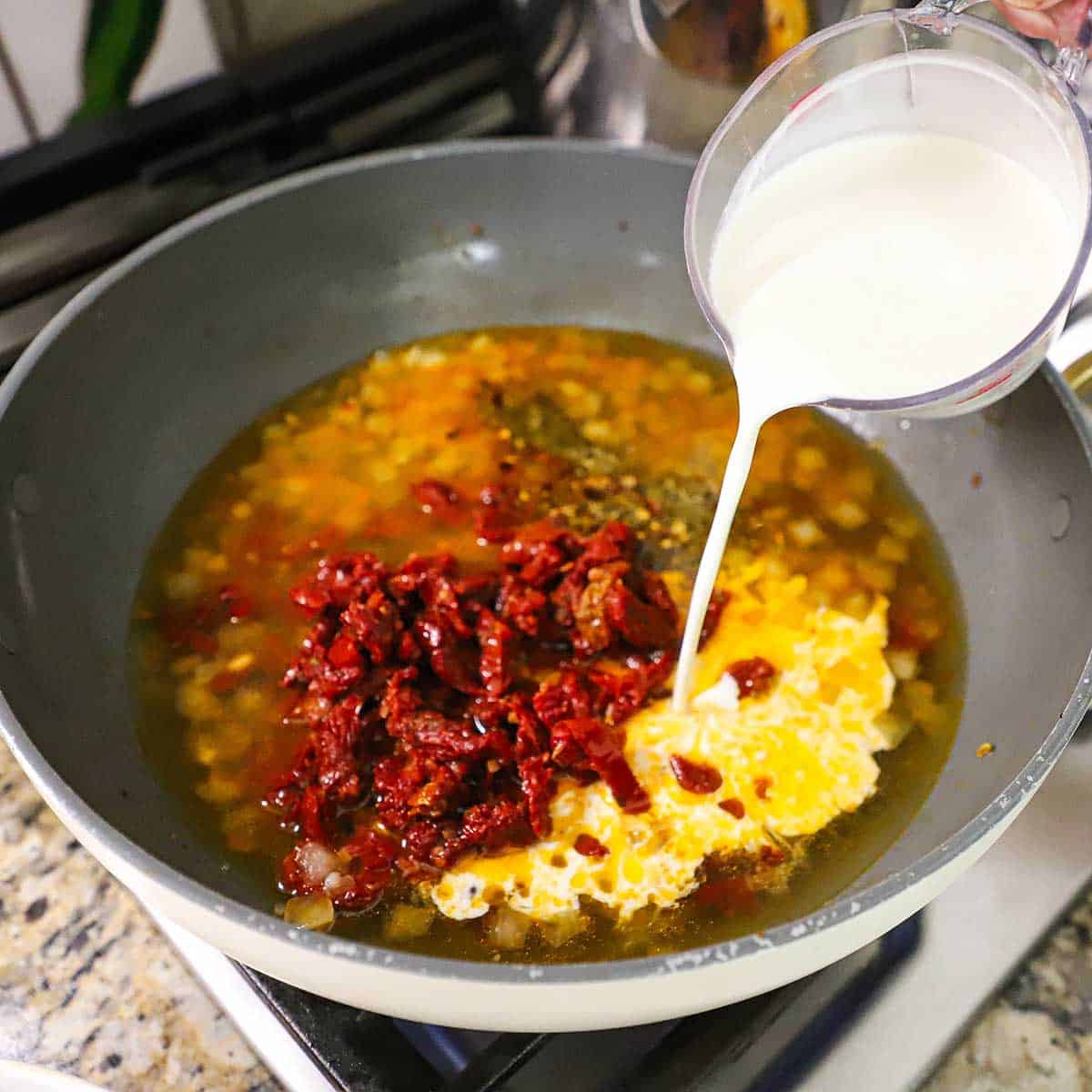 A person pouring heavy cream from a measuring cup into a large skillet filled with simmering chicken stock, strips of sun-dried tomatoes, seasonings, and sautéed onions.