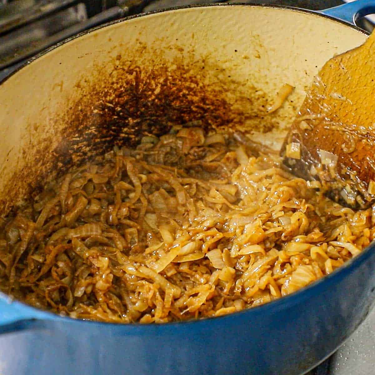 A close-up view of caramelized onions that are simmering in a large oval Dutch oven on a gas stove.