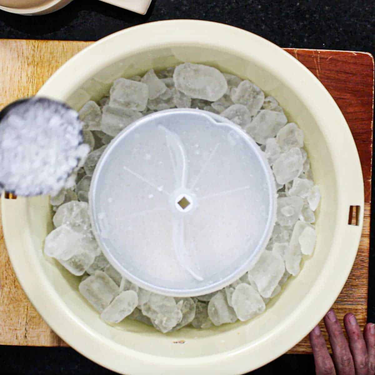 A person using a small measuring cup to transfer rock salt on ice that is circulating around the exterior of a metal canister that is churning homemade ice cream.