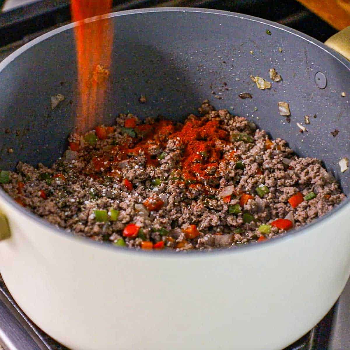 A person adding smoked paprika to cooked ground beef along with sautéd peppers and other seasonings in a large pot on a gas stove.