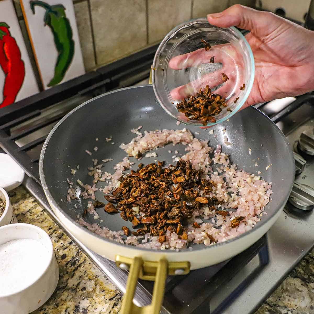 A person adding chopped porcini mushrooms from a small glass bowl into a large skillet on a gas stove containing simmering saut&eacute;ed chopped shallots and minced garlic.