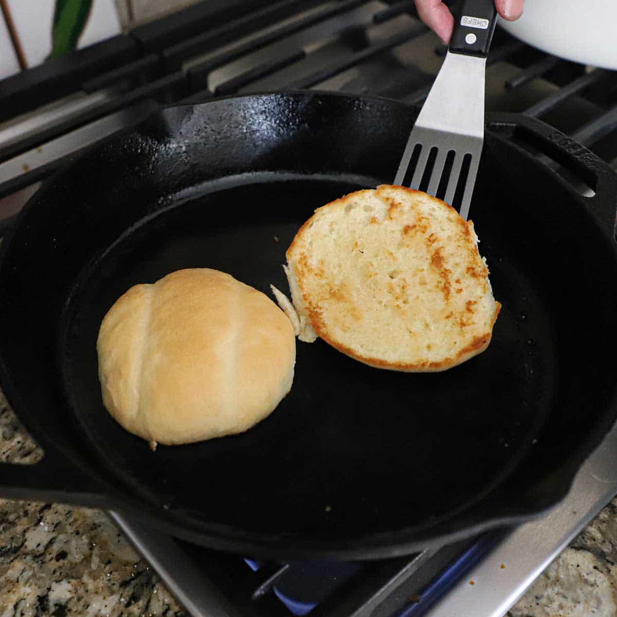 A person using a small metal spatula to flip over a toasty telera bun that has been split open and being toasted with butter on a large cast-iron skillet on a gas stove.